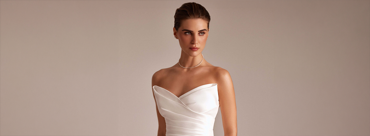A woman modeling a strapless white wedding dress with a sweetheart neckline and gathered ruching, photographed against a plain warm beige studio background.