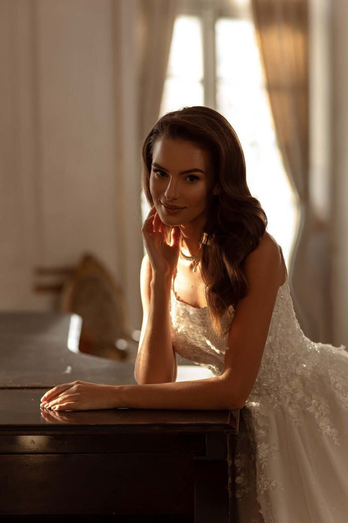 Elegant model wearing a lace wedding dress with square neckline, posing at a table with soft lighting.