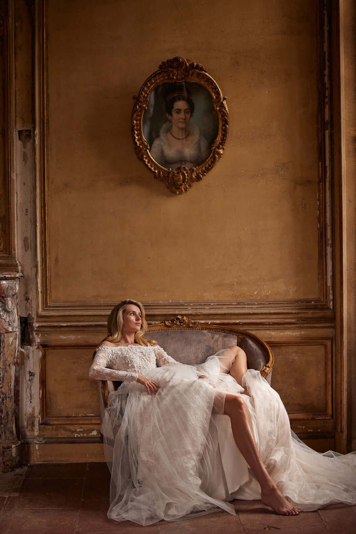 A model in a lace wedding gown lounges on a vintage sofa with an elegant backdrop featuring a classic portrait.
