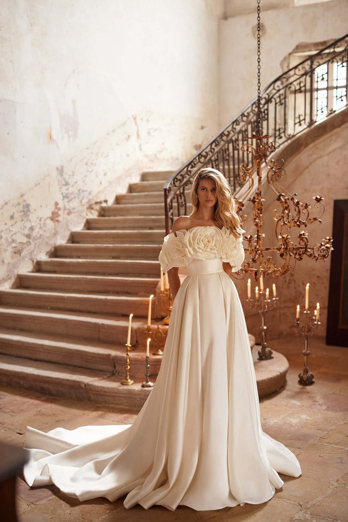 Ivory A-line Margery plain satin wedding dress with corset, open shoulders, and pleated skirt, styled in elegant setting.